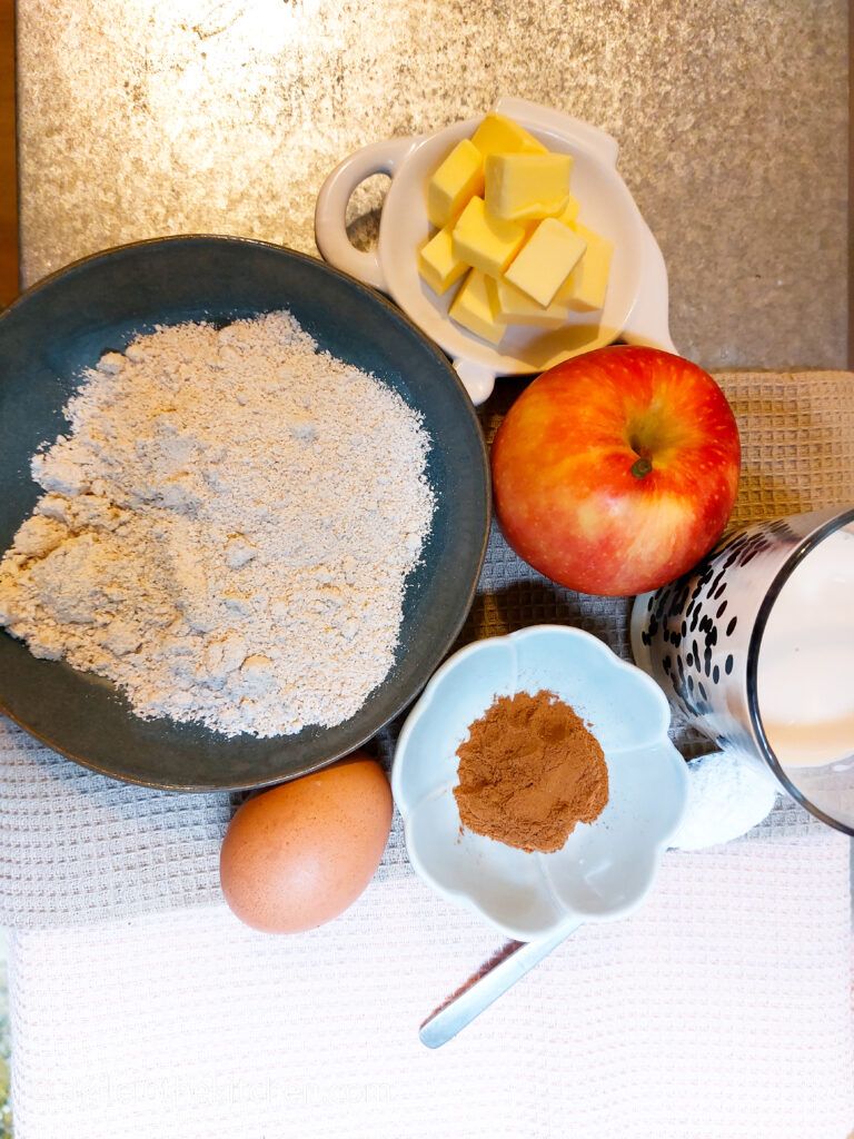 Ingredients laid out on a table to make apple cinnamon pancakes, butter diced, oat flour, red apple, oat milk, cinnamon, egg and salt