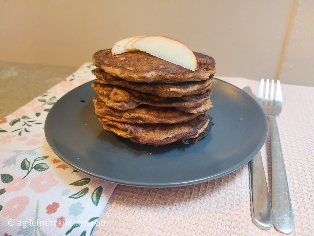 On a pink textured background, a table cloth with flowers, a black matte plate a stack of apple cinnamon pancake with slices of red apples on top, to the side a knife and fork.