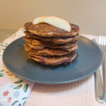 On a pink textured background, a table cloth with flowers, a black matte plate a stack of apple cinnamon pancake with slices of red apples on top, to the side a knife and fork.