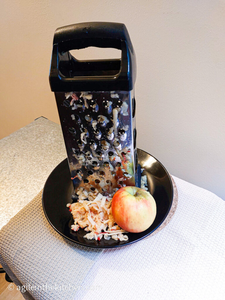 On a black plate, a standing grater, with some grated apple and an untouched apple.