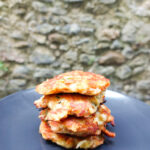 In the foreground a dark grey matte plate with a stack of four halloumi and vegetable patties.