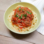 On a beige textured background,a white cloth napkin, a green bowl with spaghetti bolognese, topped with fresh basil