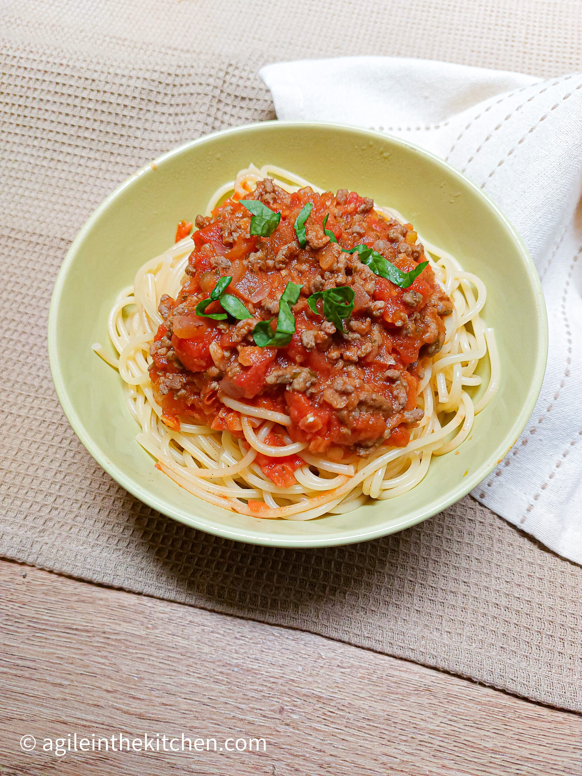 On a beige textured background,a white cloth napkin, a green bowl with spaghetti bolognese, topped with fresh basil