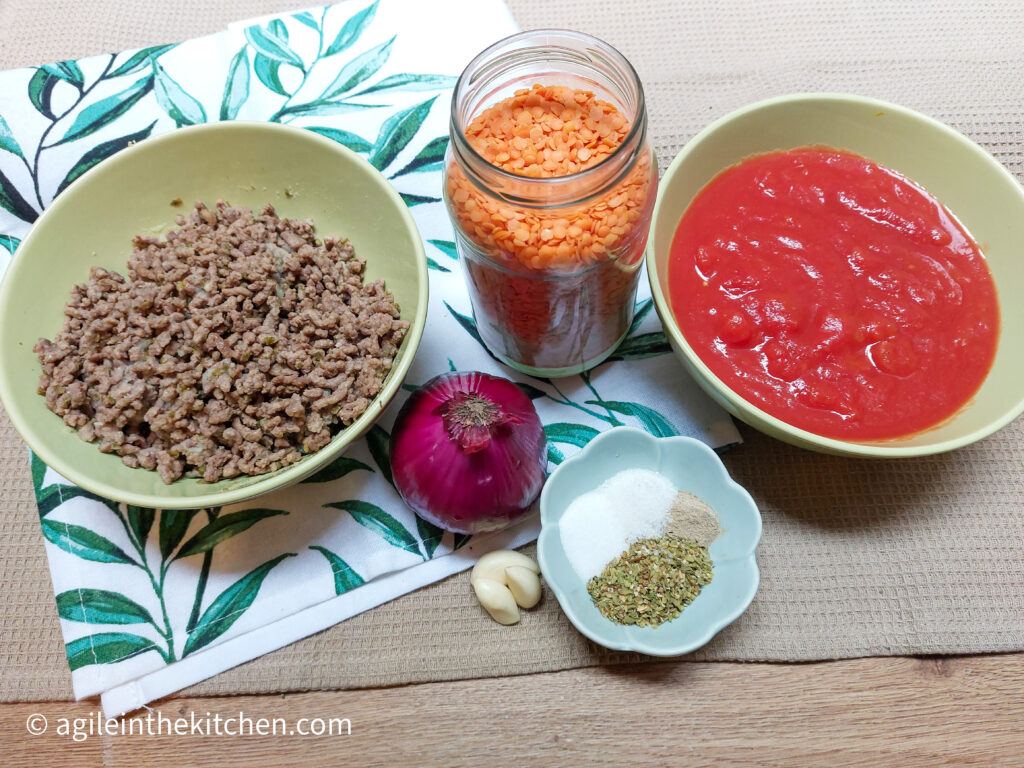 On a beige textured background, a folded cloth napkin with green leaves. On top, the ingredients to make bolognese, a green bowl with cooked mince meat, a red onion, garlic cloves, red lentils in a glass jar, a green bowl with crushed tomatoes and a small bowl with salt, sugar, oregano and white pepper.