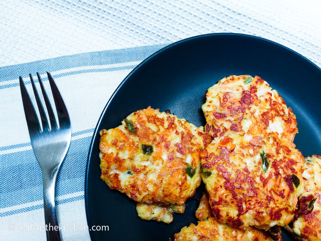 On a white textured background, a close up of a black, matte plate with halloumi and vegetable patties, next to a fork and underneath the plate and fork, folded blue striped cloth napkin