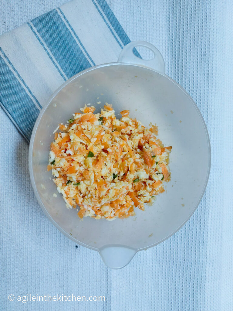 On a white background, a white and blue striped cloth napkin folded. On top of the napkin a plastic bowl of halloumi and vegetable patty mizx, shredded