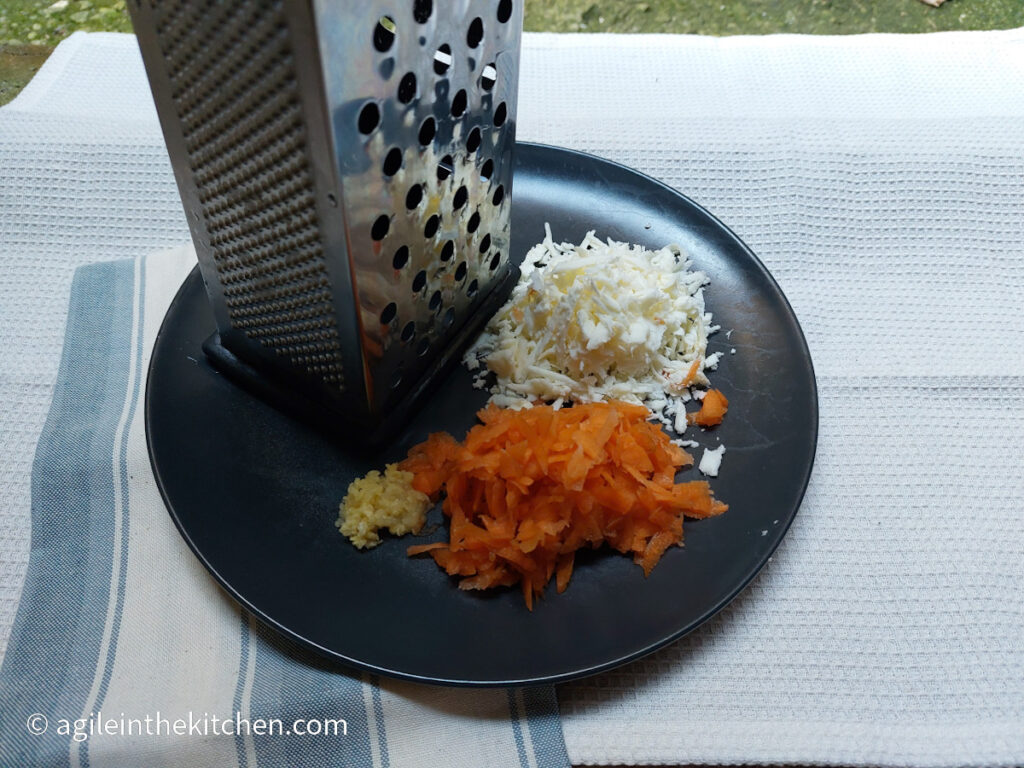 On a white background a blue and white striped cloth napkin. On top of the napkin a black plate with a hand held vegetable shredder and a larger pile of shredded halloumi, next to a large pile of shredded carrots and a small pile of shredded garlic.