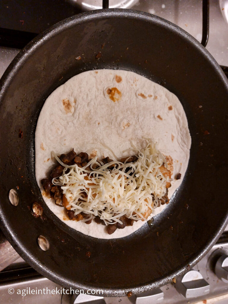 In a frying pan, an open tortilla bread with black beans, scampi and shredded cheese, ready to be folded into a quesadilla