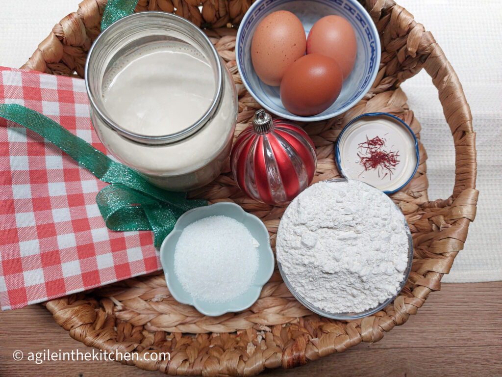 In a wicker tray the ingredients to make saffron pancakes, oat milk, three eggs, a pinch of saffron, flour and sugar. Together with a folded red, gingham cloth napkin, a strand of green string and a red striped Christmas bauble.