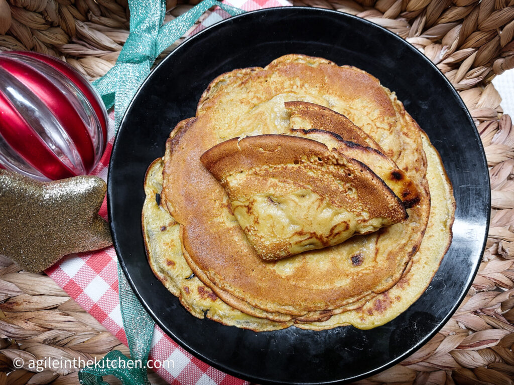 In wicker basket with a striped Christmas bauble, a golden star, a black plate with saffron pancakes piled up with the top one folded in fours.