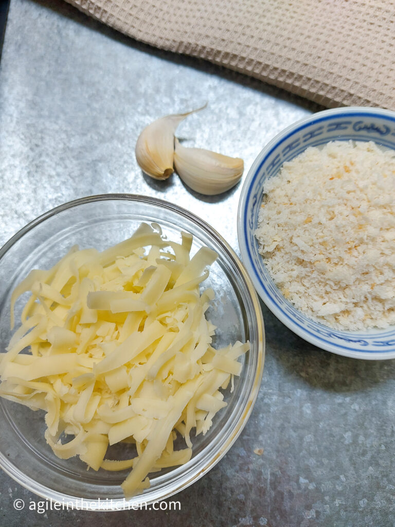 On a silver background, a glass bowl with shredded cheese, two cloves of garlic and a bowl of panko bread crumbs