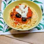 On a beige textured background a white cloth napkin with green leaves, a green bowl with spaghetti and Mediterranean tomato sauce with feta cheese and black olives on top.