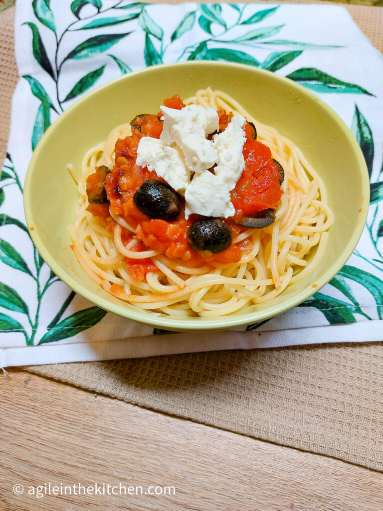 On a beige textured background a white cloth napkin with green leaves, a green bowl with spaghetti and Mediterranean tomato sauce with feta cheese and black olives on top.