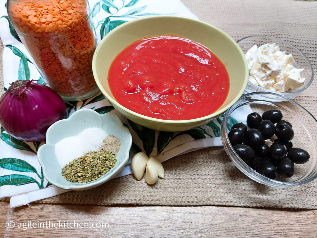 On a beige textured background the ingredients to make Mediterranean tomato sauce, one red onion, split red lentils, a green bowl with crushed tomatoes, a bowl of feta cheese, a bowl of black olives, three garlic cloves and a pot of herbs salt, sugar, oregano and black pepper.