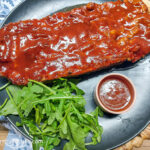 On a textured background, a black matte plate with a barbecue meatloaf, with a handful of green leaves and a smaller pink bowl of barbecue sauce.