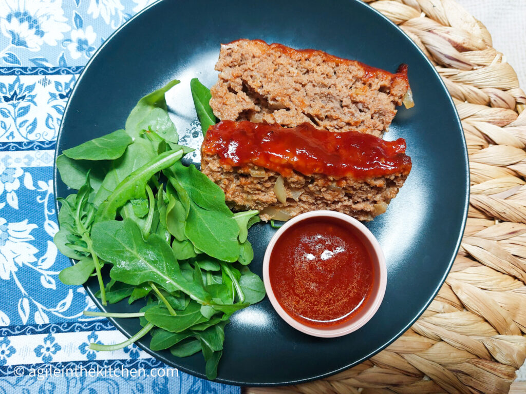 On a textured background with a blue flower patterned table cloth on the left side, in the middle a black matte plate, with two slices of barbecue meatloaf, a handful of green leaves and a smaller pink bowl with barbecue sauce.