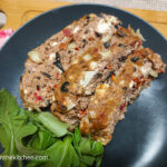 On a wooden cutting board with a red gingham cloth in the left corner, a black matte plate with three slices of Mediterranean meatloaf and a handful of green leaves.