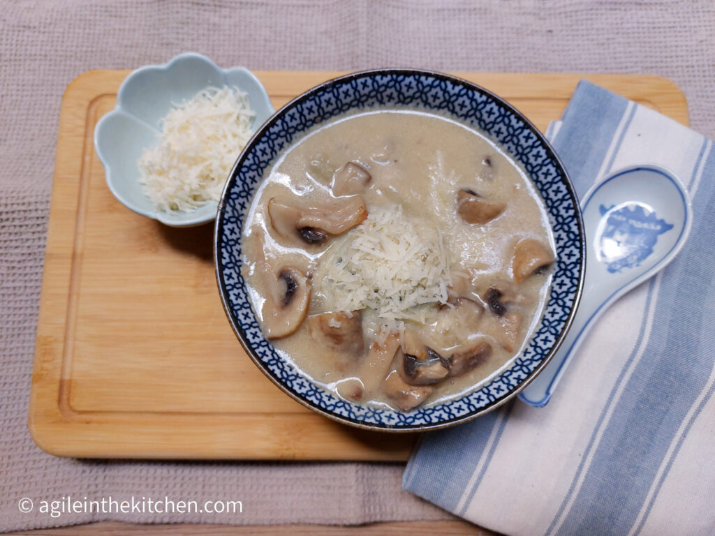 On a wooden cutting board a blue striped cloth napkin to the left of the picture with a porcelain spoon on top, a blue patterned soup bowl with mushroom soup, topped with shredded Parmesan cheese. In the left upper corner, a flower shaped bowl with shredded Parmesan cheese