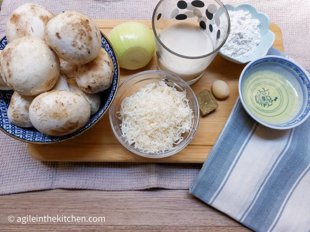 On a wooden cutting board, the ingredients to make creamy mushroom soup, from top left: a bowl of mushrooms, one yellow onion, a glass bowl of shredded Parmesan cheese, a glass of cream, one bouillon cube, one clove of garlic, a flower shaped bowl with flower, a bowl with oil. In the right lower corner a blue and white striped folded cloth napkin.