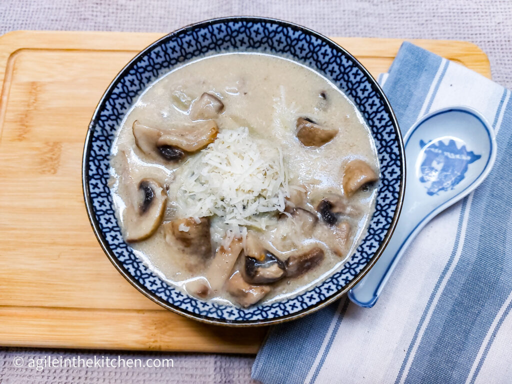 On a wooden cutting board a blue striped cloth napkin to the left of the picture with a porcelain spoon on top, a blue patterned soup bowl with mushroom soup, topped with shredded Parmesan cheese.