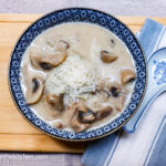 On a wooden cutting board a blue striped cloth napkin to the left of the picture with a porcelain spoon on top, a blue patterned soup bowl with mushroom soup, topped with shredded Parmesan cheese.