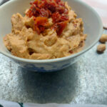 On a silver background, a bowl of sun-dried tomatoes hummus with extra sun-dried tomatoes chopped up on top.