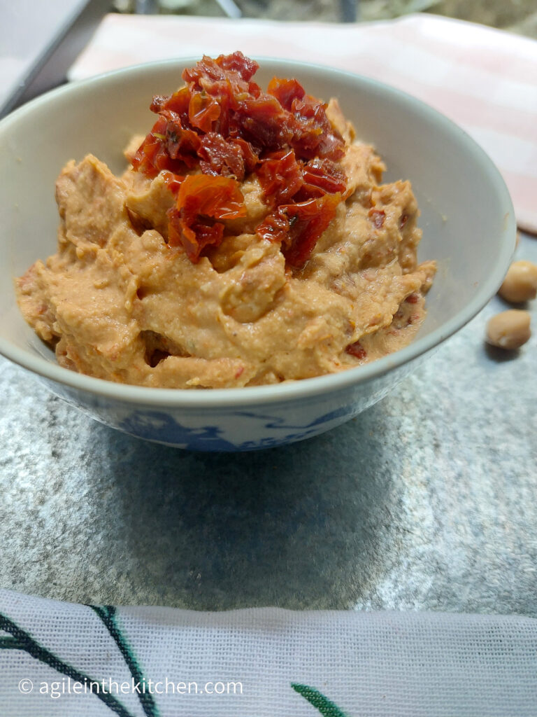 On a silver background, a bowl of sun-dried tomatoes hummus with extra sun-dried tomatoes chopped up on top.