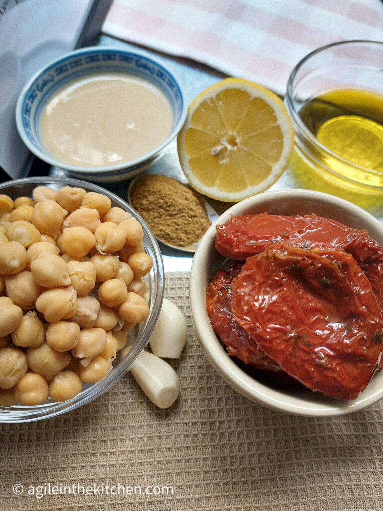 On a light brown textured background, the ingredients to make sun-dried tomatoes hummus. Clockwise: in a bowl, tahini, on a teaspoon cumin, half a lemon, in a glass bowl olive oil, in a cup sun-dried tomatoes, two cloves of garlic, a glass bowl of chickpeas.