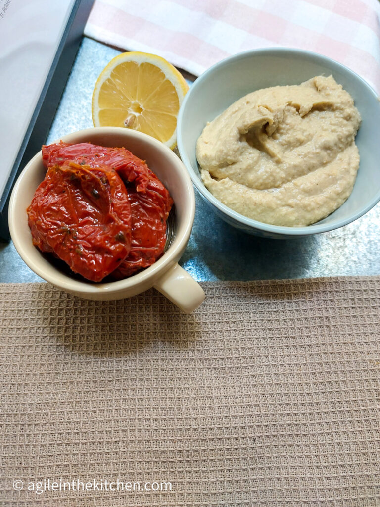 On a silver background with a brown textured table cloth, a cup of sun dried tomatoes, half of a lemon and a bowl of basic hummus