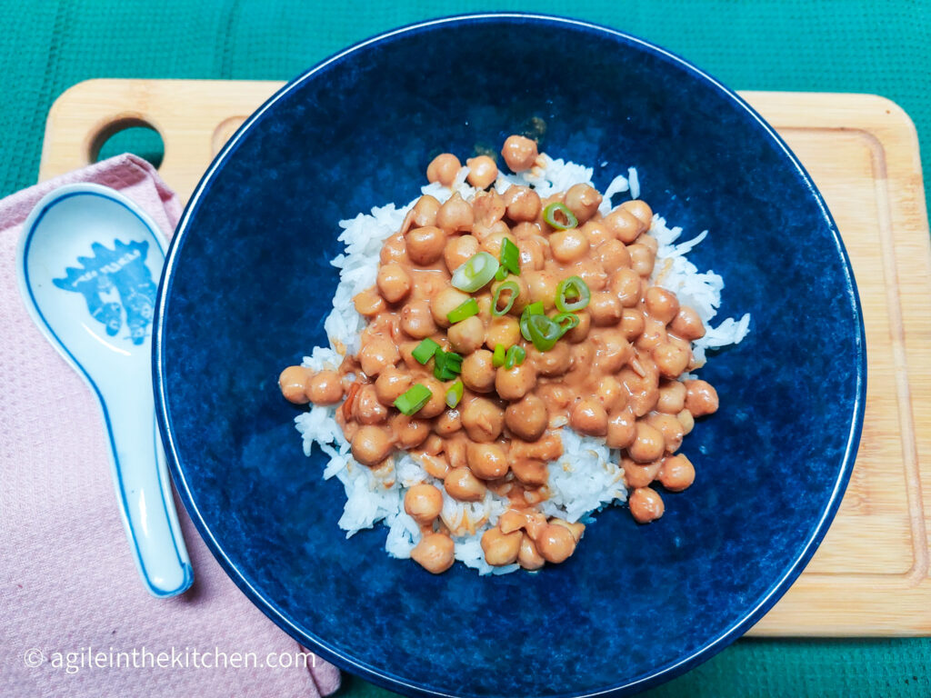 A wooden cutting board on top of a green textured background, in the left corner a pink cloth napkin with a ceramic spoon on top. In the middle a blue bowl with rice, chickpea peanut butter curry and a sprinkling of green onions.