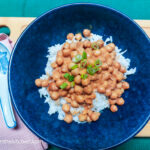 A wooden cutting board on top of a green textured background, in the left corner a pink cloth napkin with a ceramic spoon on top. In the middle a blue bowl with rice, chickpea peanut butter curry and a sprinkling of green onions.