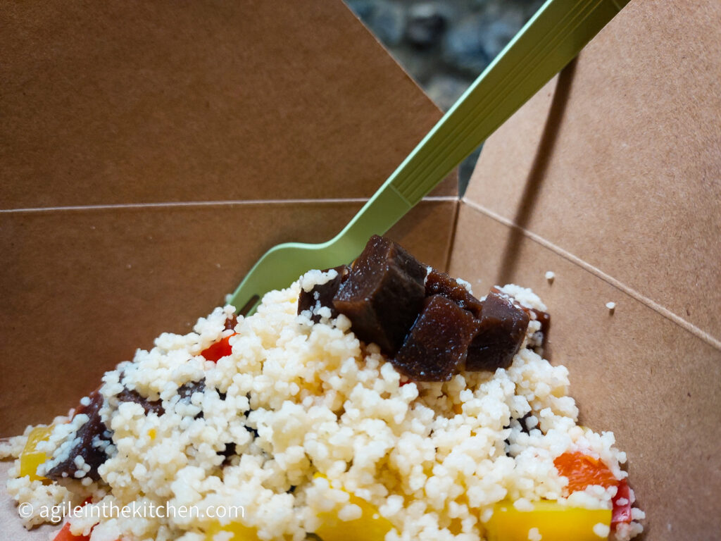 In a brown paper box, with a green plastic fork sticking out of it, a couscous salad with pickled vegetables, pieces of beetroot sprinkled on top.