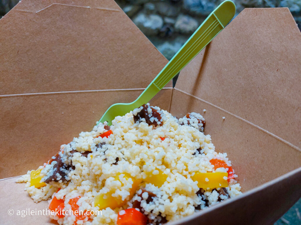 In a brown paper box, with a green plastic fork sticking out of it, a couscous salad with pickled vegetables, pieces of beetroot, yellow pepper and red pepper visible.