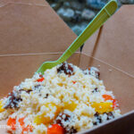 In a brown paper box, with a green plastic fork sticking out of it, a couscous salad with pickled vegetables, pieces of beetroot, yellow pepper and red pepper visible.