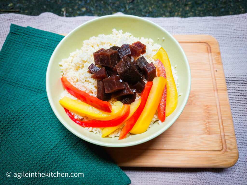 On a beige textured background, a wooden cutting board with a green folded cloth napkin, a green bowl with couscous, red and yellow pickled peppers and a pile of diced pickled beetroot