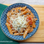 On a cutting board, draped with a green textured cloth napkin, a blue patterned bowl with vegetarian cheeseburger pasta, topped with shredded cheese.