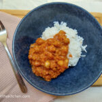 On a beige textured background, a wooden cutting board, mostly covered in a pink, textured cloth napkin with a fork on top and a blue bowl with basmati rice and chickpea stroganoff.