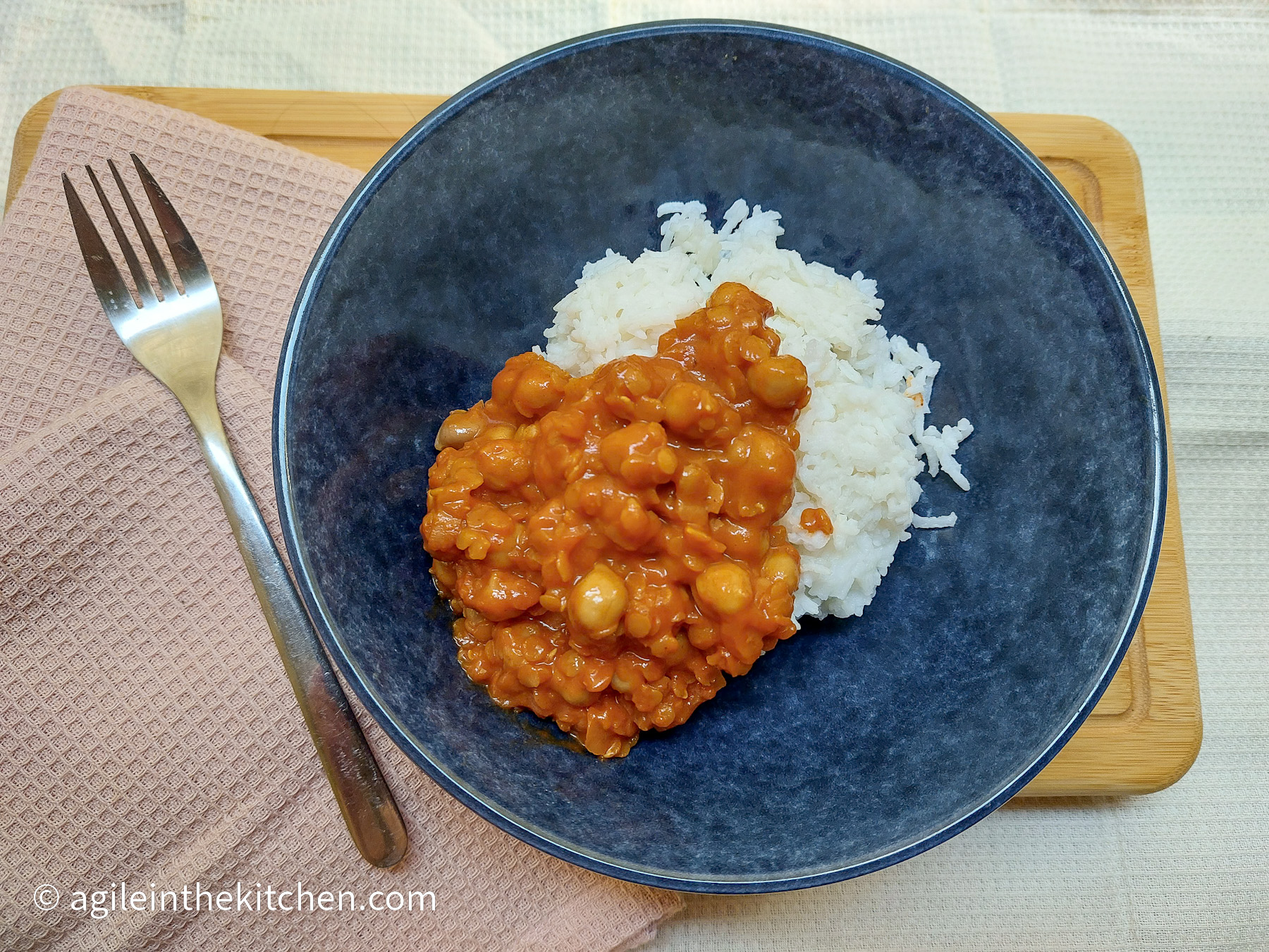 On a beige textured background, a wooden cutting board, mostly covered in a pink, textured cloth napkin with a fork on top and a blue bowl with basmati rice and chickpea stroganoff.