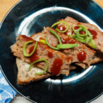 On a cutting board, with a blue, flower patterned cloth napkin in the left corner. In the middle, a black plate with a sourdough open faced sandwich with three slices of barbecue meatloaf, on top a few drops of barbecue sauce, sprinkled with a few strands on green onion.