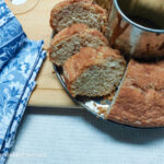 A close up of, a white background, a wooden cutting board and to the left part of a blue flower patterned cloth napkin. To the right, part of a circular cake tin with a cinnamon cake, three slices have been made in the cake and are turned to see the inside of the cake.