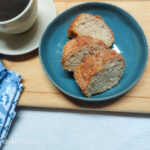 On a white textured background, a wooden cutting board with a blue, flower patterned cloth napkin in the lower left corner. In the upper left corner, the edge of a white coffee cup with black coffee. In the middle of the photo, a blue plate with three slices of cinnamon cake.