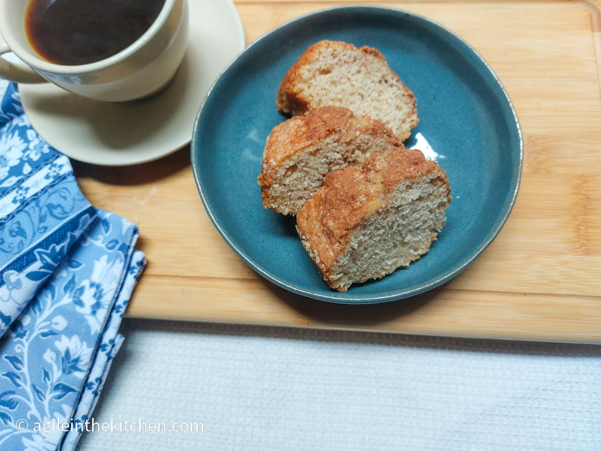 On a white textured background, a wooden cutting board with a blue, flower patterned cloth napkin in the lower left corner. In the upper left corner, the edge of a white coffee cup with black coffee. In the middle of the photo, a blue plate with three slices of cinnamon cake.