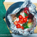 On a white background, on a wooden cutting board with a green cloth napkin to the left, a black matte plate with a foil package with fish, shrimp, halved cocktail tomatoes and spinach leaves.