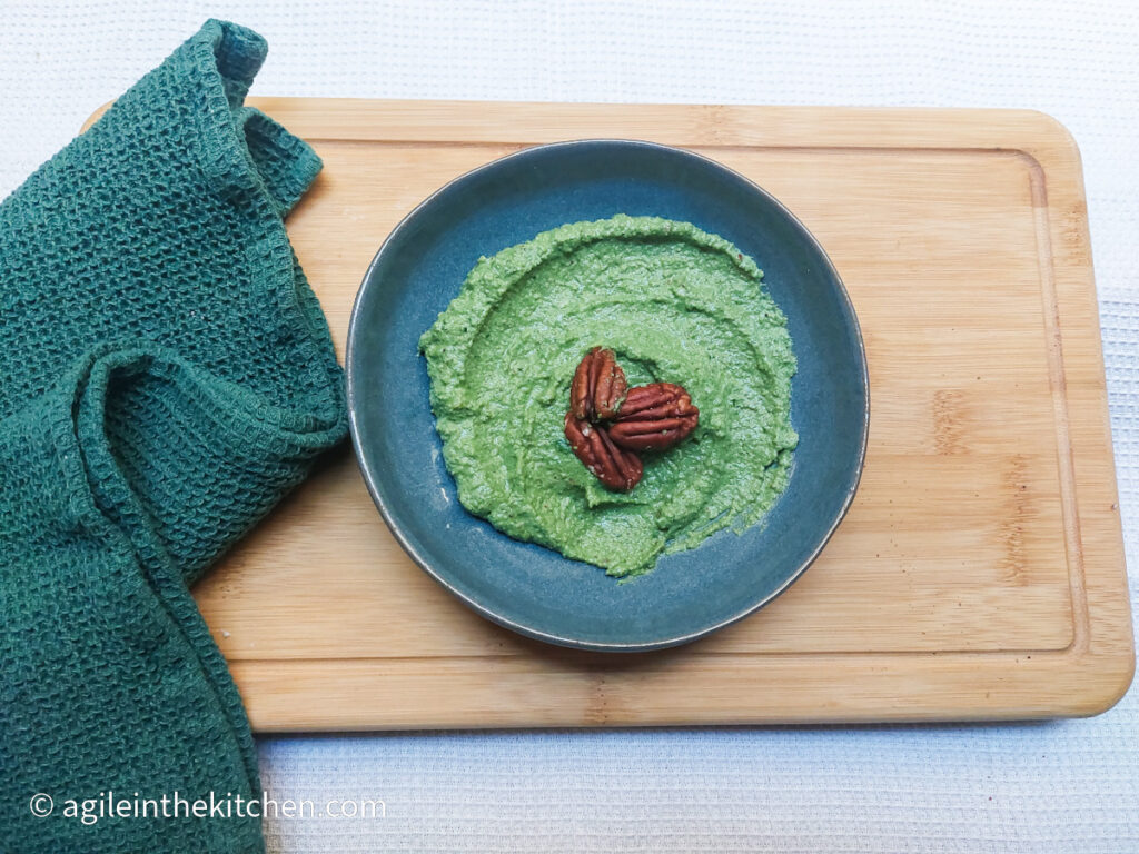 On a white background, on a wooden cutting board with a green cloth napkin folded to the left. In the middle, a blue plate with green spinach pesto topped with three pecan nuts.