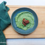 On a white background, on a wooden cutting board with a green cloth napkin folded to the left. In the middle, a blue plate with green spinach pesto topped with three pecan nuts.