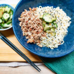 On a wooden cutting board, a green cloth napkin in the right corner, in the middle a larger blue bowl with noodles, shredded teriyaki tofu, topped with spicy cucumber salad and black sesame seeds. Further to the left, a smaller bowl with spicy cucumber salad. Below this bowl, a pair of black chopsticks.