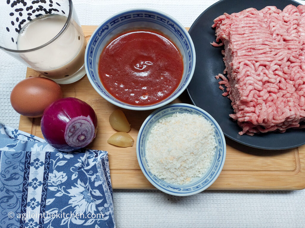 On a white background, a wooden cutting board, in the left hand lower corner a blue patterned cloth napkin. Clockwise from top left, ingredients to make barbecue meatballs; a glass of milk, a bowl of barbecue sauce, a plate with minced meat, a bowl of panko bread crumbs, two cloves of garlic, one red onion and one egg.