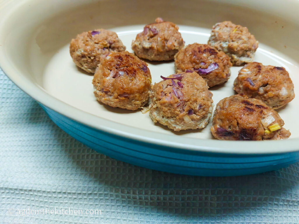 On a white, textured back ground, a blue, oval oven form with eight barbecue meatballs.
