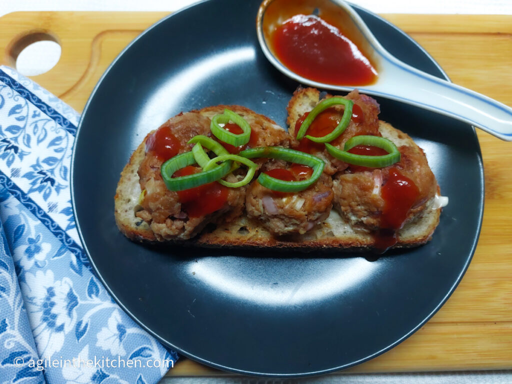 On a white background, a wooden cutting board, in the left hand lower corner a blue patterned cloth napkin. In the middle of the photo, a black, matte plate with a meatball sandwich on sourdough, with barbecue sauce and green onion on top and a porcelain spoon with the rest of the barbecue sauce.