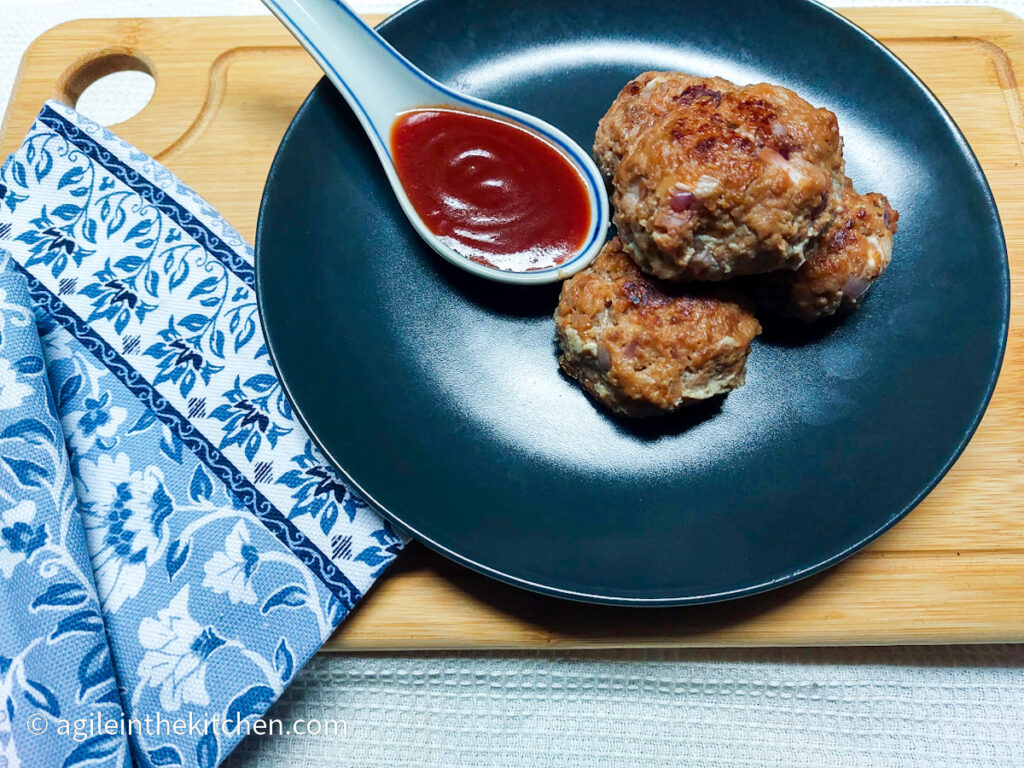 On a white background, a wooden cutting board, in the left hand lower corner a blue patterned cloth napkin. In the middle of the photo, a black, matte plate with a stash of meatballs, and a porcelain spoon with barbecue sauce.